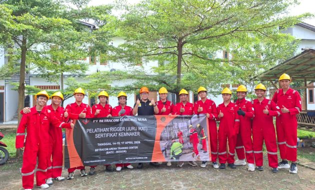 Group of trainees in red coveralls and yellow hard hats posing outdoors, holding a banner for a training program.