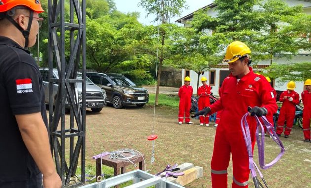 Outdoor safety training: crew in red coveralls and yellow helmets monitor a lifting drill as a concrete weight is lowered into a metal-framed pit beneath a crane structure.